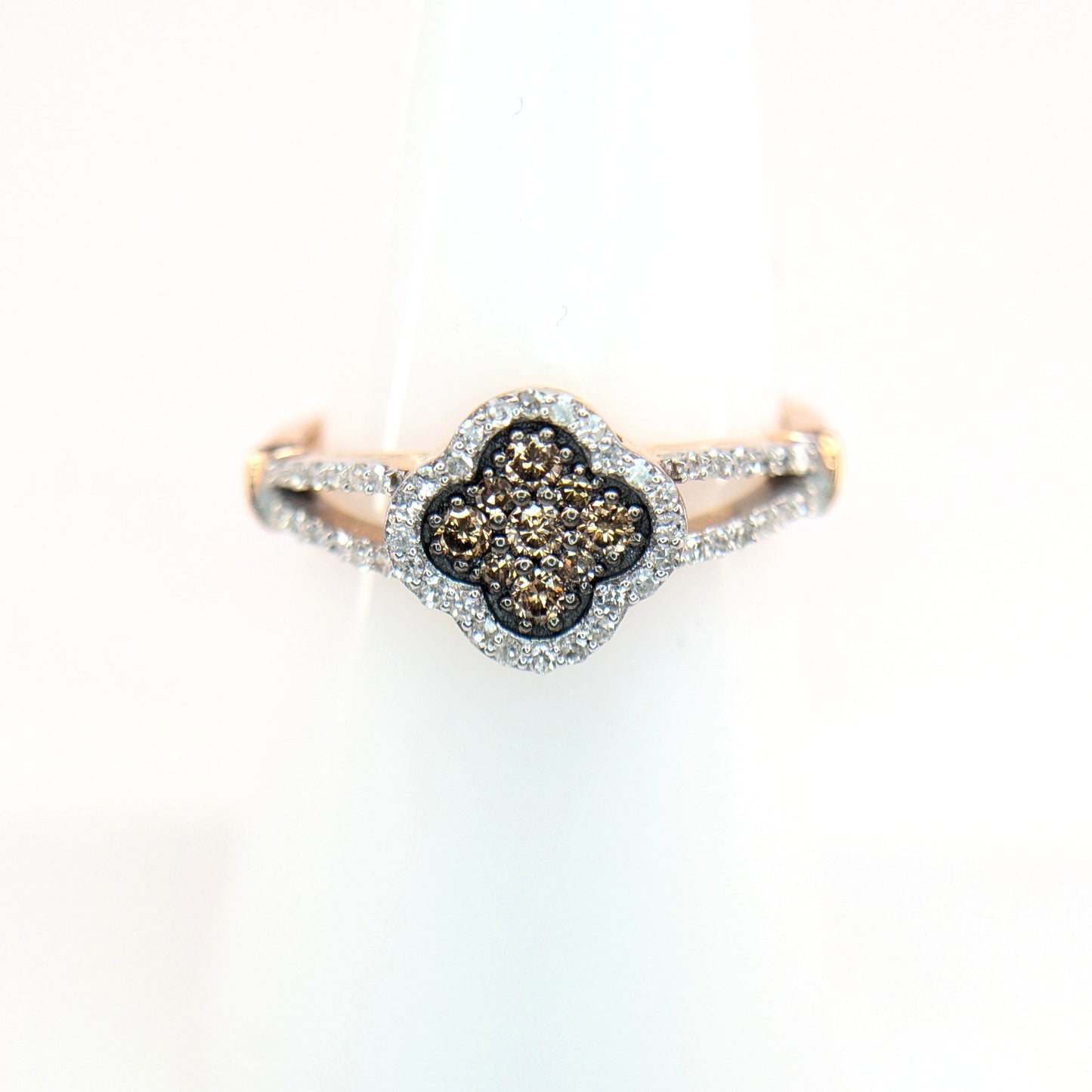 A rose gold ring with four-leaf clover. The center of the ring contains cognac diamond with an outer layer or white diamonds. The Ring sparkles on a white display stand.