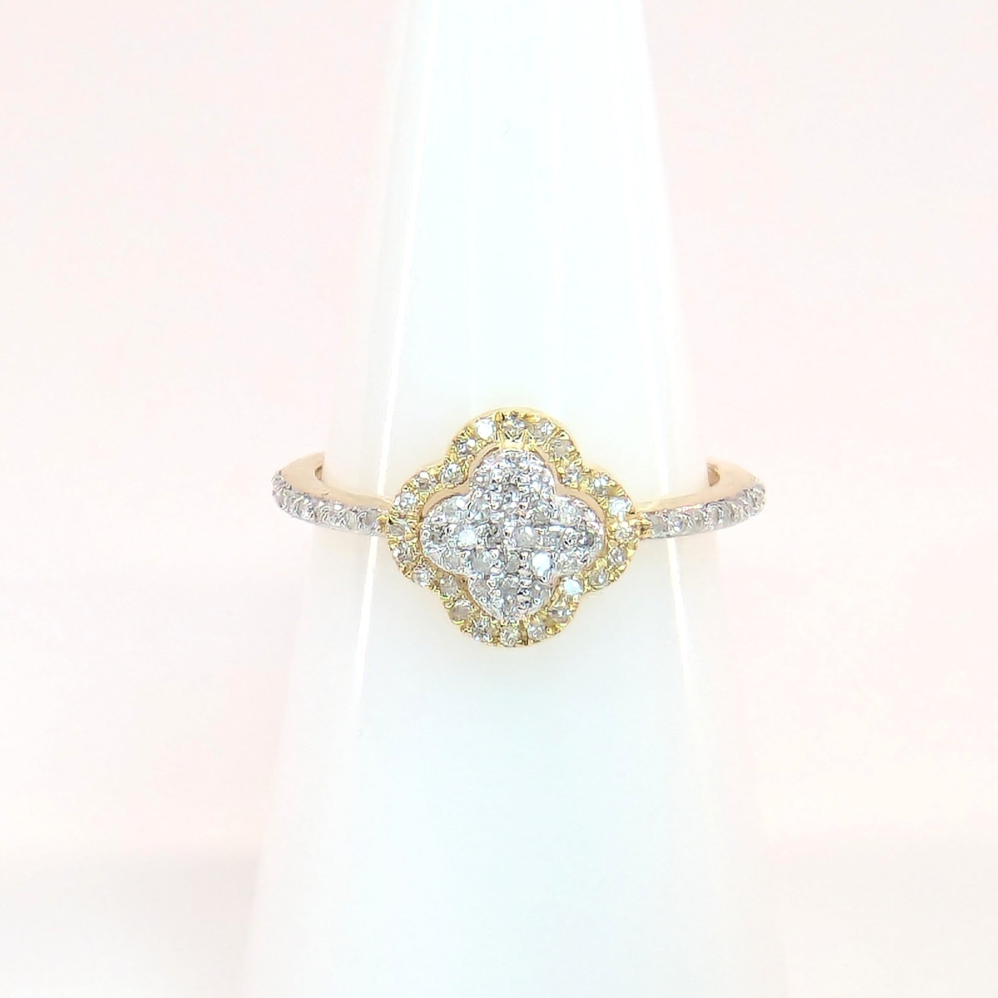 Close-up shows a sparkling diamond yellow gold ring featuring a clover design resting on a white display stand.
